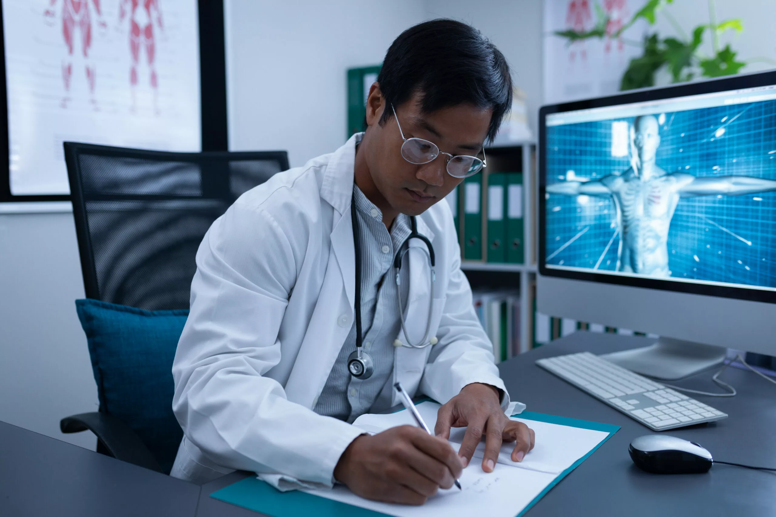 South Asian Dr in a lab coat writing at his desk with a computer in the background