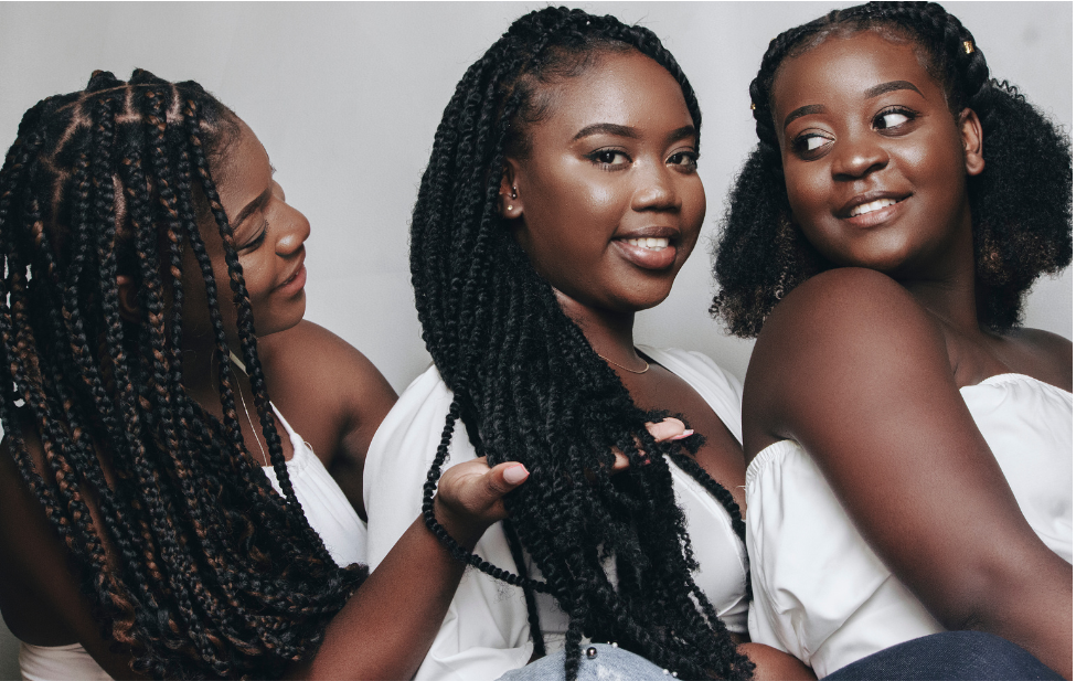 three Black women in white clothes stroking each other's braided hair